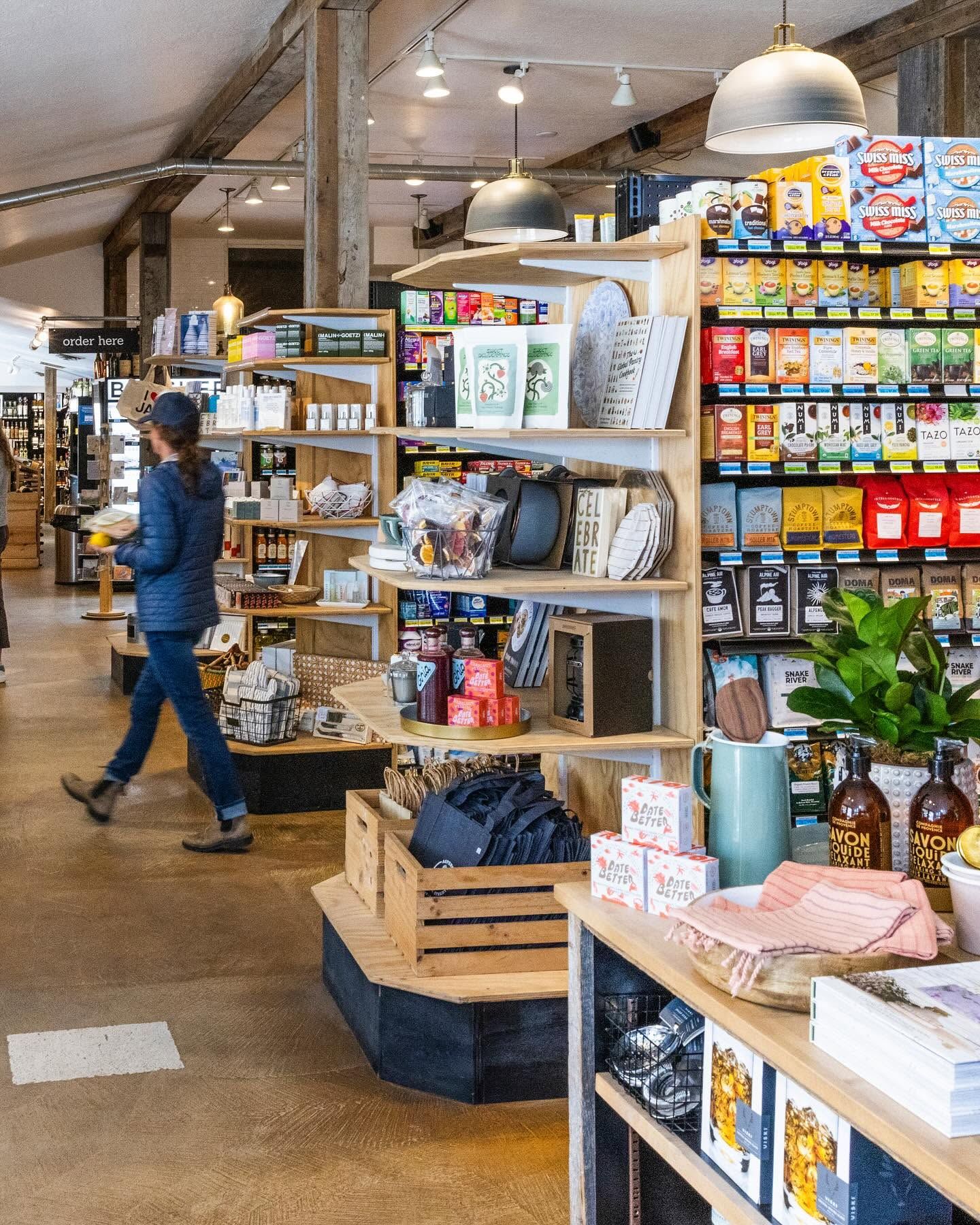 A shopper walks past well-stocked wooden shelves inside Pearl Street Market, browsing an eclectic mix of specialty teas, coffee beans, cookbooks, home goods, and local pantry items. Warm pendant lighting and exposed beams give the market a bright, welcoming feel. 