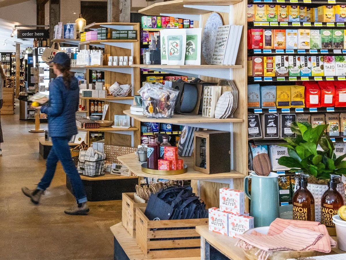 A shopper walks past well-stocked wooden shelves inside Pearl Street Market, browsing an eclectic mix of specialty teas, coffee beans, cookbooks, home goods, and local pantry items. Warm pendant lighting and exposed beams give the market a bright, welcoming feel.