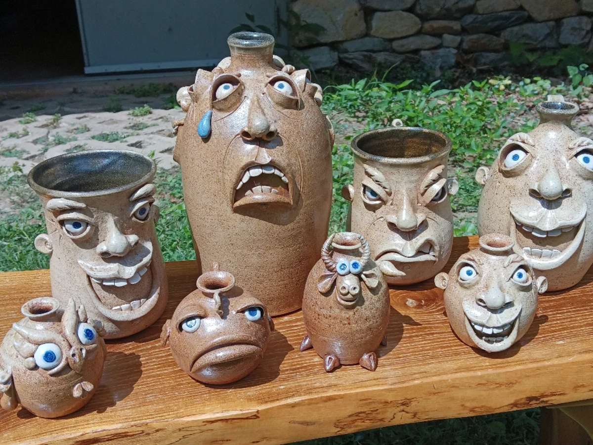 A collection of whimsical pottery jugs and vases with exaggerated facial expressions and cartoonish features sits on a wooden bench outdoors.
