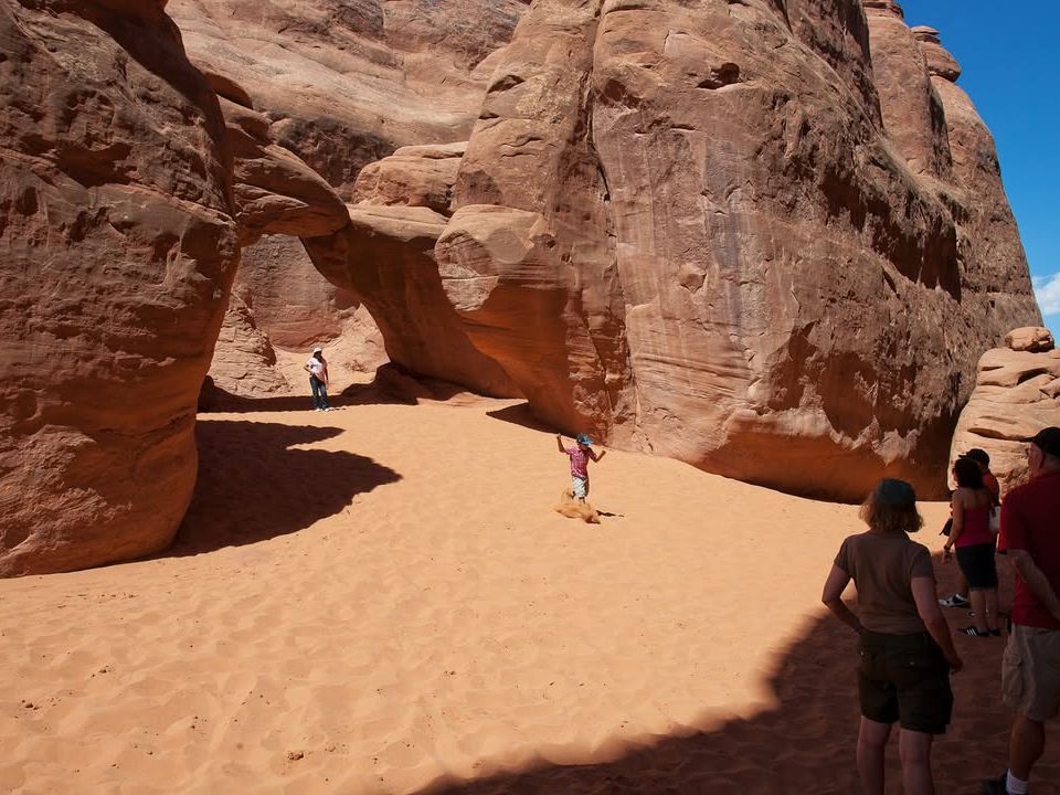 Tourists explore a dramatic sandstone arch at Arches National Park, with red rock formations towering under clear blue skies. The soft desert sand and natural shadows offer an adventurous yet peaceful hike.