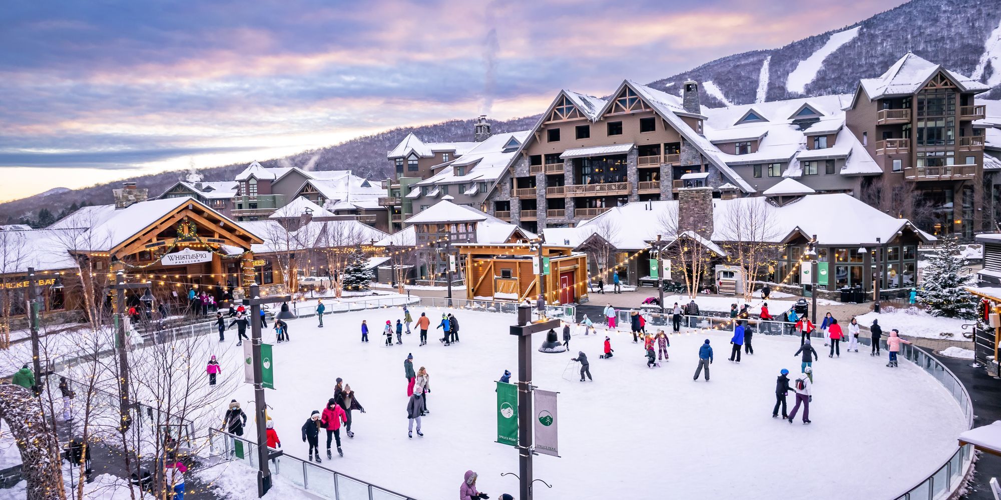 Families skate across an outdoor ice rink in Stowe, Vermont, surrounded by alpine lodges twinkling with winter lights. Snowy slopes rise in the background, creating a festive mountain-town atmosphere.  File Name: Stowe | Vacation Rentals