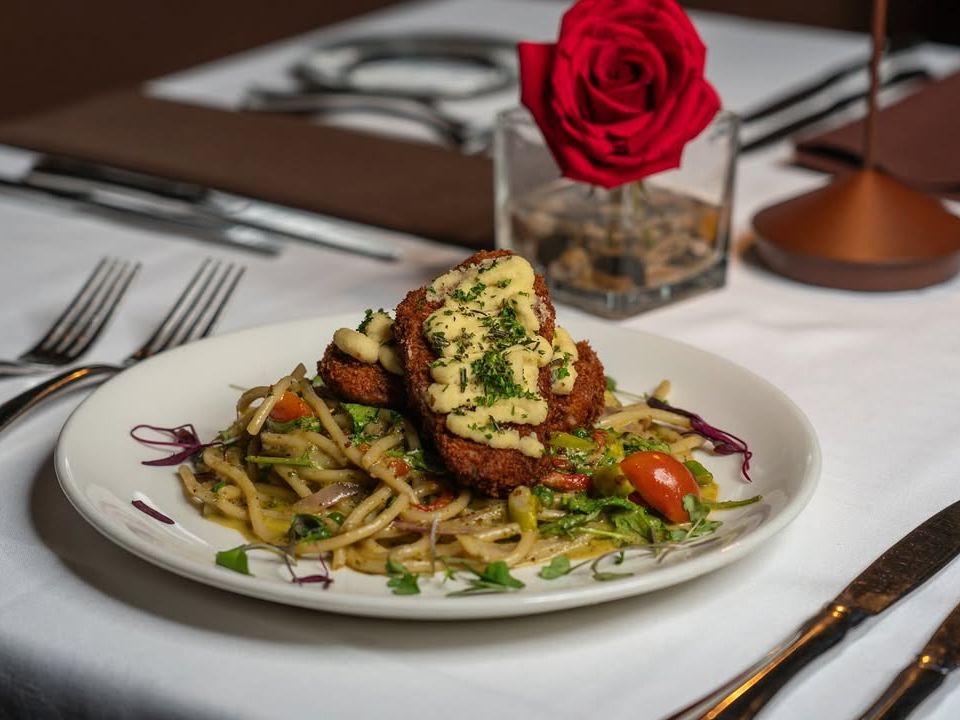 A fine dining plate from Kres Chophouse featuring breaded meat topped with creamy sauce over a bed of pasta and vegetables. Elegant presentation in a romantic setting.