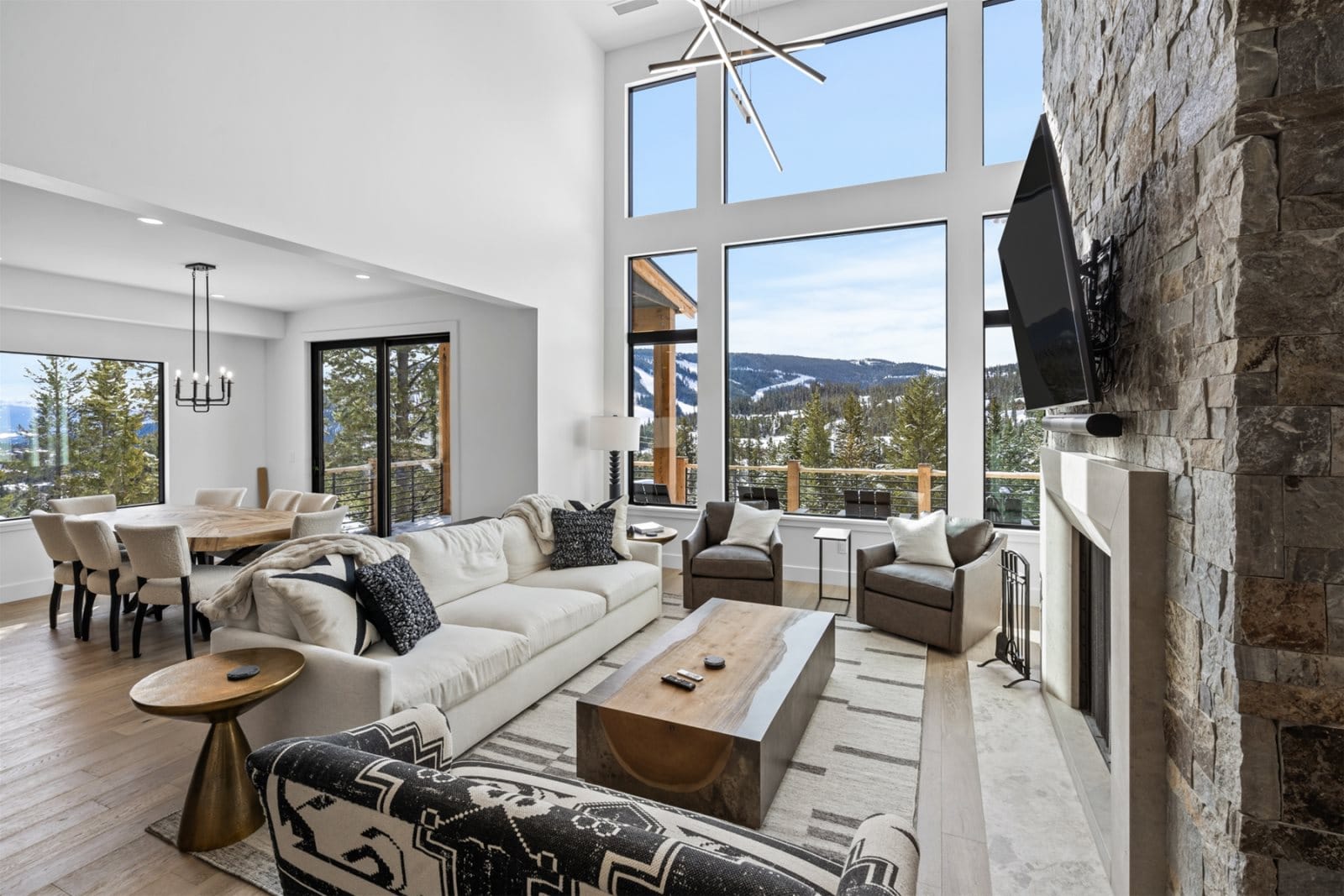 A bright mountain great room with floor-to-ceiling windows showcasing snowy forest views, a stone fireplace with mounted TV, neutral seating, and an open dining area filled with natural light.