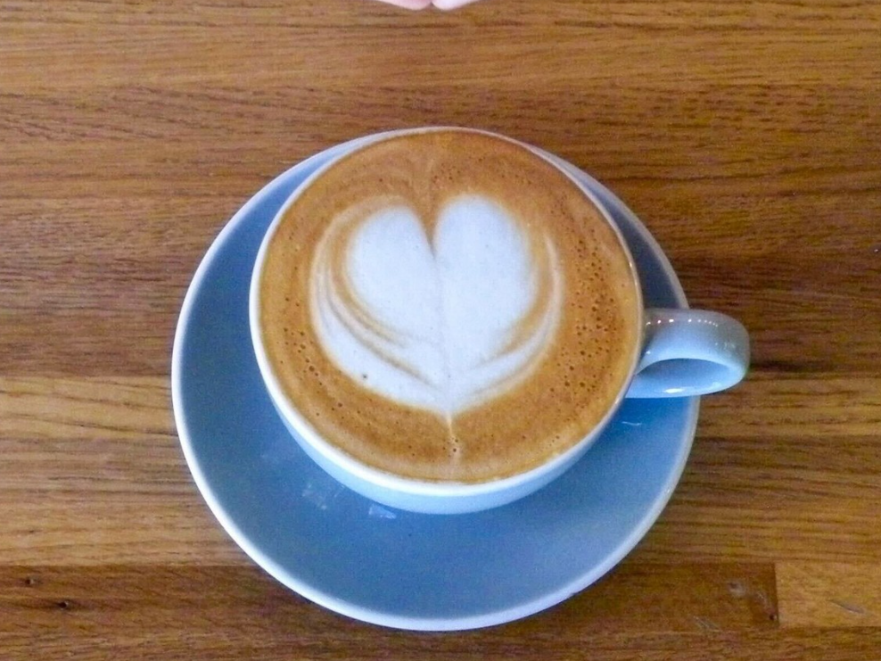 A cup of cappuccino with a heart-shaped latte art design on its frothy surface, served in a light blue ceramic cup and saucer on a wooden table.
