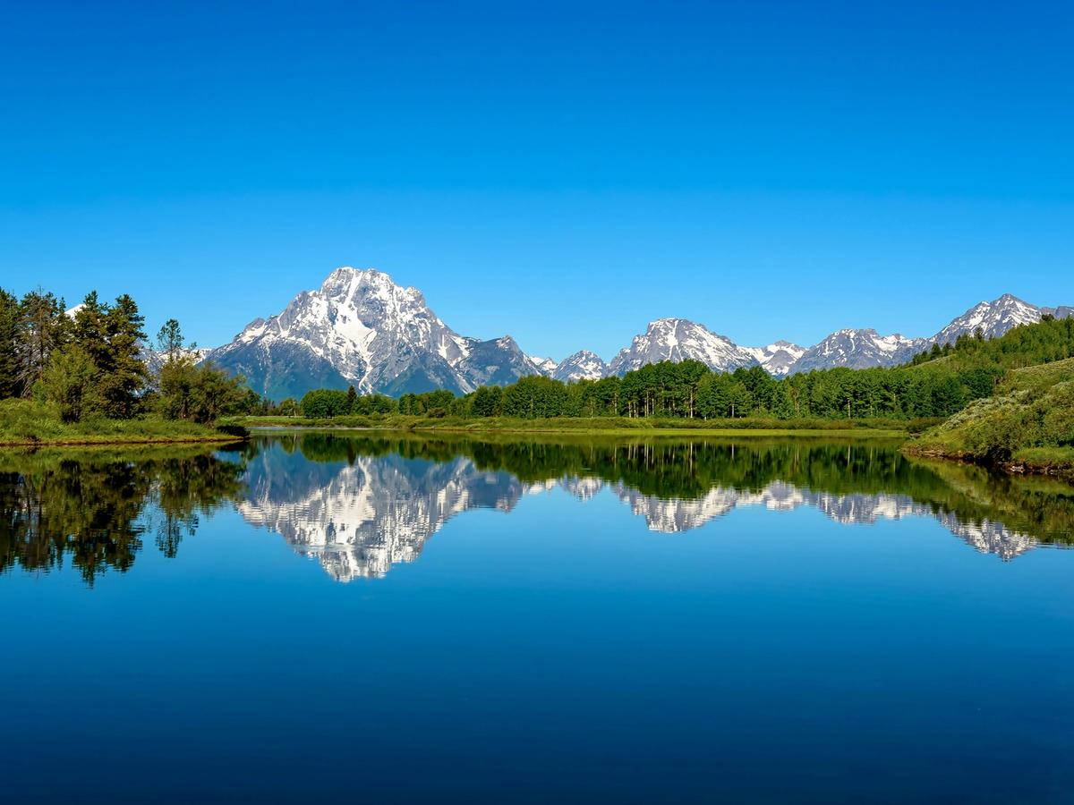 Snow-capped peaks of the Teton Range are perfectly mirrored in the still, glassy waters of a lake, framed by lush green trees and a brilliant blue sky.