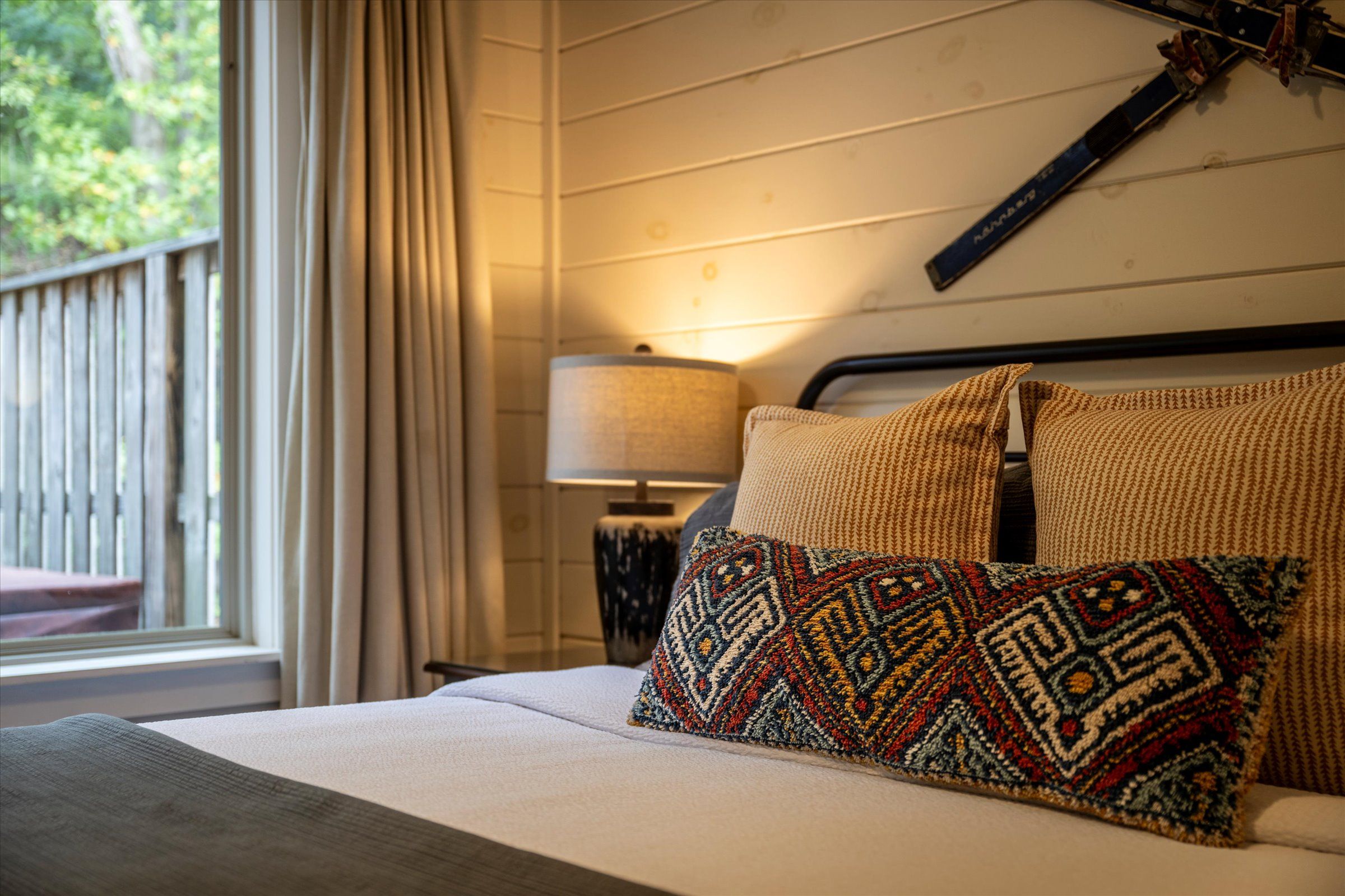 A cozy bedroom with a rustic vibe, featuring a textured throw pillow on a neatly made bed, warm lighting from a bedside lamp, wood-paneled walls, and vintage skis mounted as decor above the headboard.