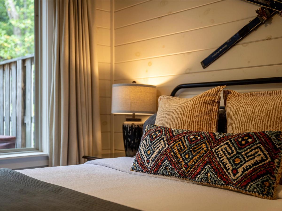 A cozy bedroom with a rustic vibe, featuring a textured throw pillow on a neatly made bed, warm lighting from a bedside lamp, wood-paneled walls, and vintage skis mounted as decor above the headboard.