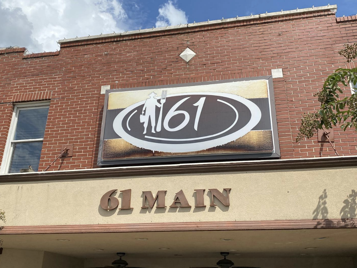 The exterior of a brick building displays a sign for "61 Main" featuring a stylized logo with a silhouette holding a fork and kettle, set against a bright blue sky with clouds.