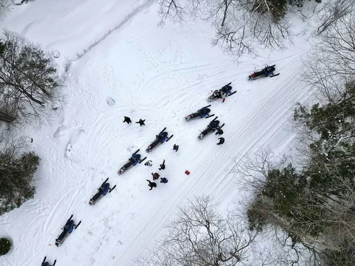 Aerial view of snowmobiles parked along a snowy forest trail, with people gathered nearby and winter trees surrounding the scene.