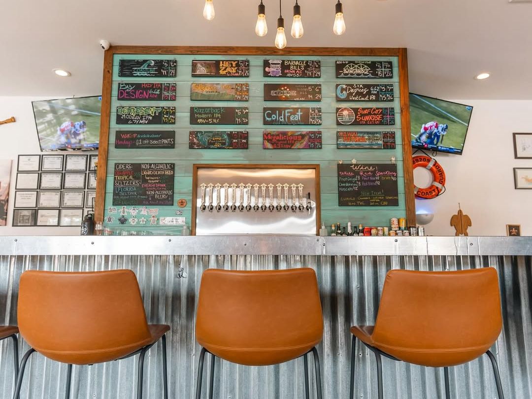 A colorful tap list hangs above a row of beer taps inside Salty Turtle Beer Co., with cozy leather stools lining the bar. This welcoming brewery is known for creative craft brews, beach vibes, and a friendly local crowd.