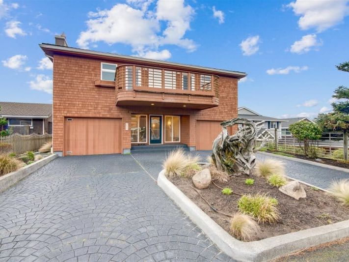 A modern coastal home featuring warm cedar siding, wide balconies, and a unique front garden sculpture. The bright driveway and open design create a relaxing atmosphere perfect for seaside getaways.