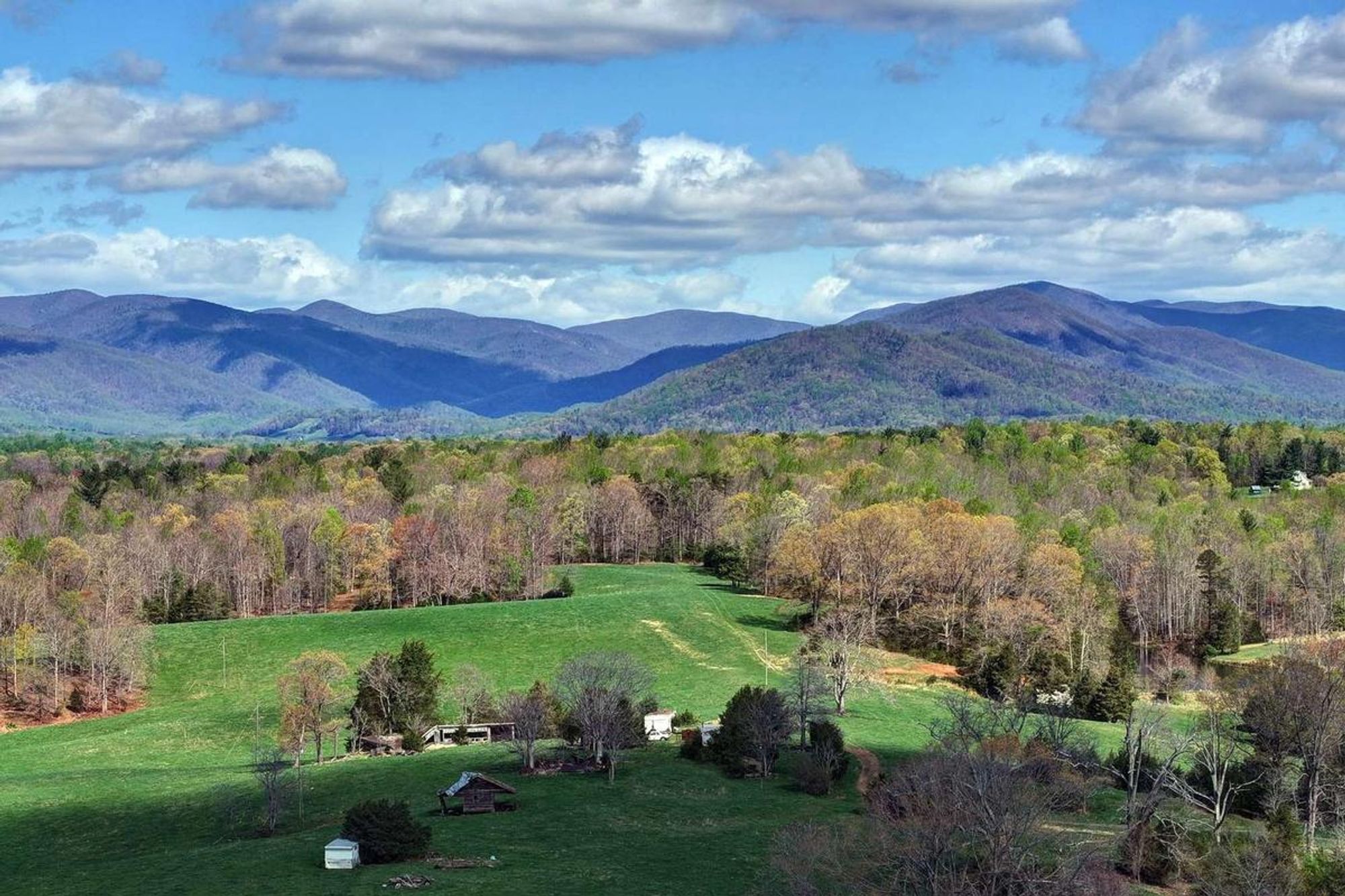 A wide open view of the Blue Ridge Mountains with soft rolling hills, lush green fields, and scattered trees in early spring bloom. Small rustic barns and cabins sit quietly at the edge of the forest, adding charm to this peaceful mountain valley.