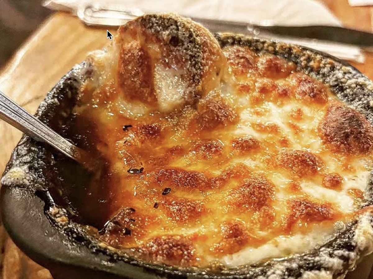 A bowl of French onion soup topped with a thick layer of melted, golden-brown cheese and a toasted bread slice, served in a rustic crock with a spoon.