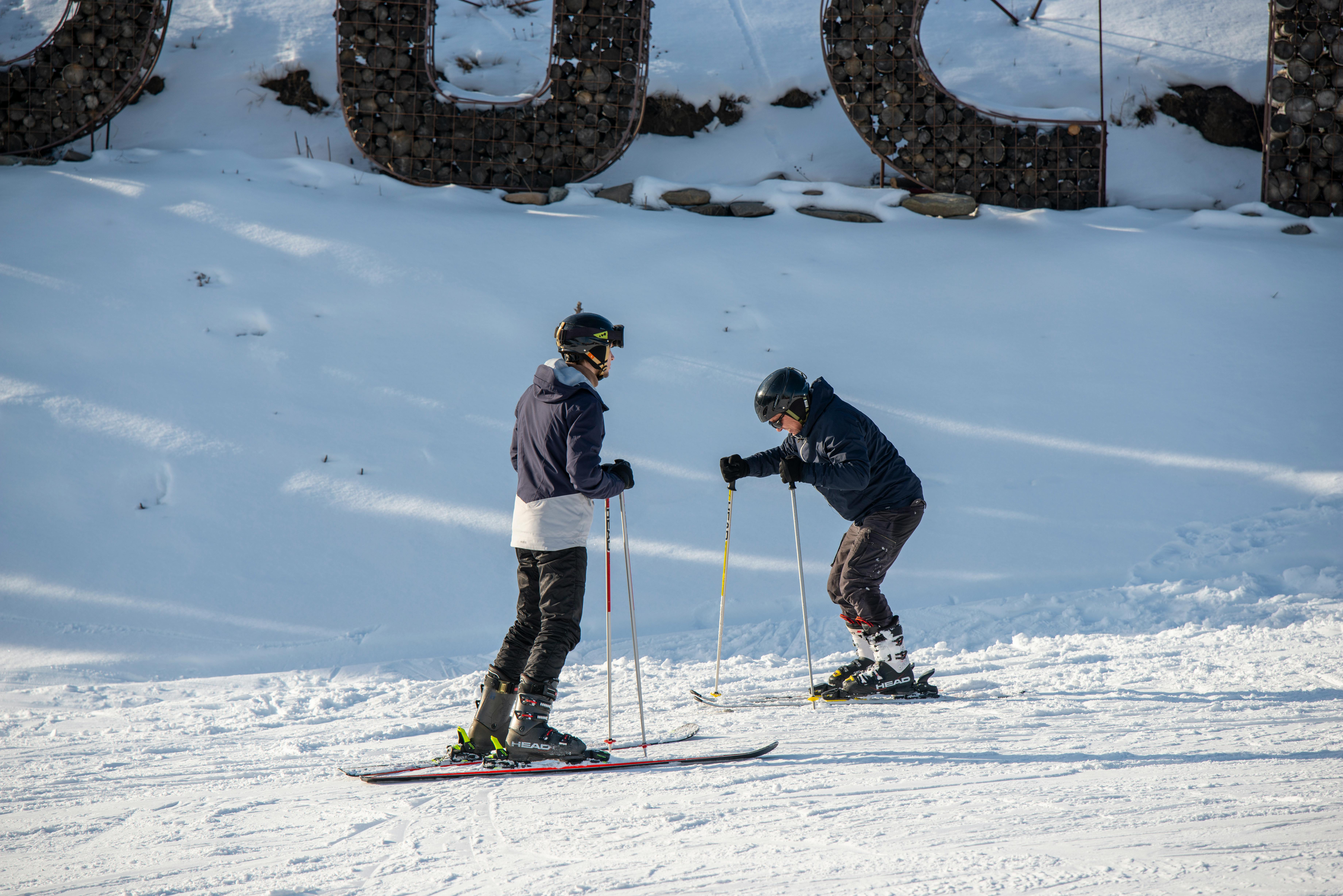 wo skiers practice on a snowy slope during a bright winter day at a ski resort. One skier leans forward with poles planted while the other stands nearby, both wearing helmets and winter gear. The fresh snow and mountain setting highlight a fun and active day of skiing outdoors.