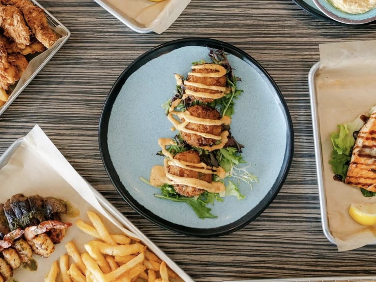 Overhead view of a seafood feast including grilled fish, fried shrimp, crab cakes with sauce, fries, hummus with crackers, and sides on a striped table.