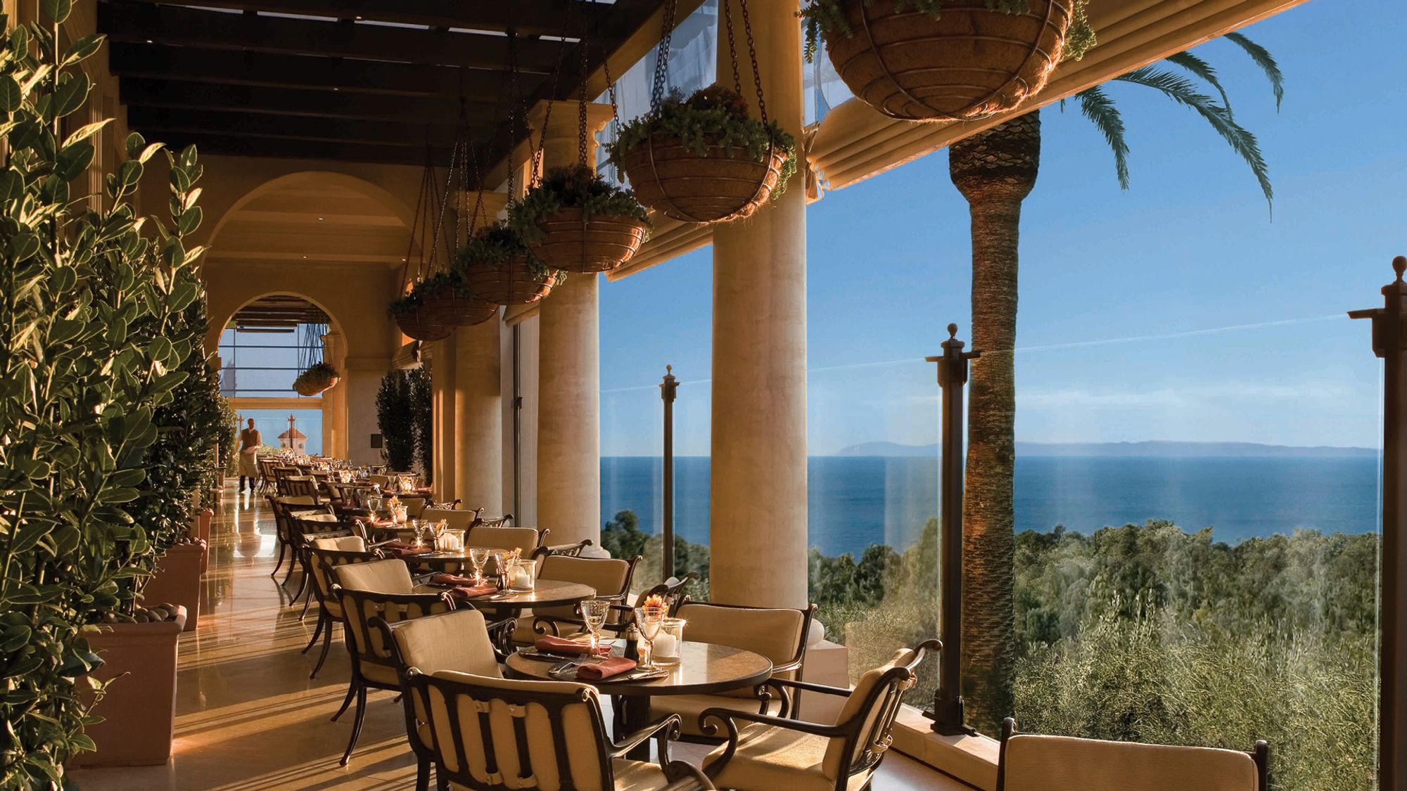 Elegant oceanview dining setup at Pelican Grill, with golden sunlight, hanging plants, and classic chairs facing the sea. A stunning spot for sunset dinners by the coast.