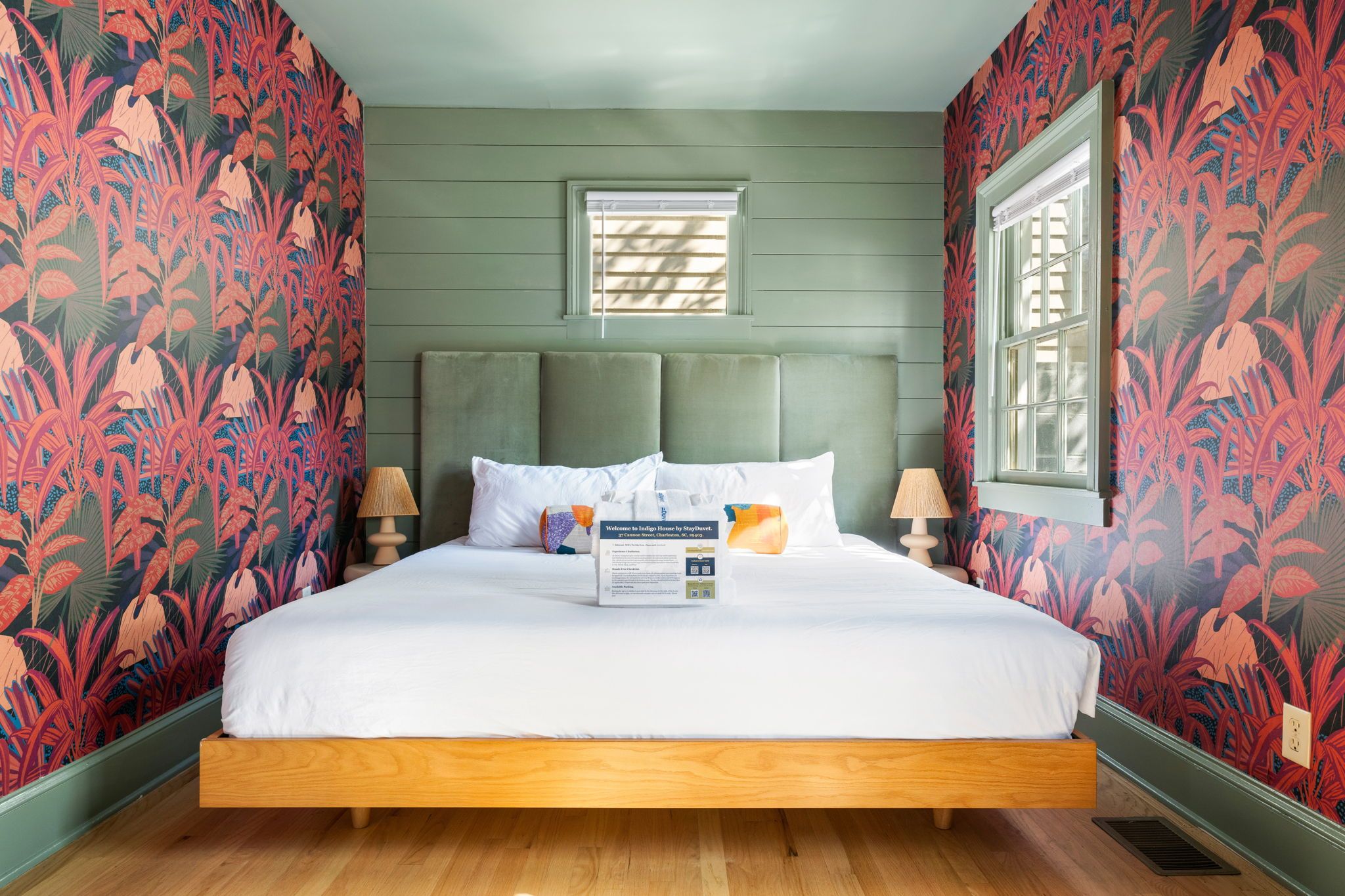 A colorful bedroom features a modern green paneled accent wall, vibrant red and pink botanical wallpaper, a platform bed with a tall velvet headboard, and two small lamps on either side.