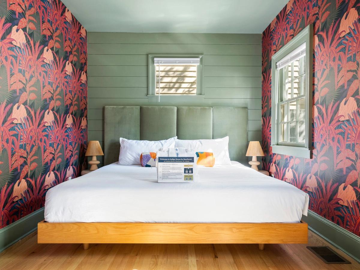 A colorful bedroom features a modern green paneled accent wall, vibrant red and pink botanical wallpaper, a platform bed with a tall velvet headboard, and two small lamps on either side.