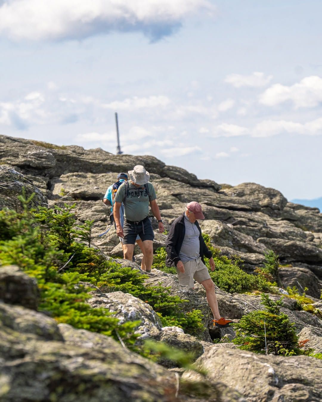 Hikers scramble over rugged granite rock formations near the summit of Mount Mansfield at Stowe Mountain Resort in Vermont, with the summit communications tower and sweeping views of the Green Mountains visible in the background on a partly cloudy summer day. 