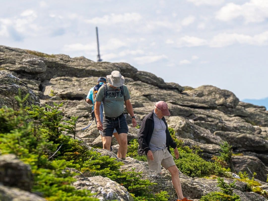 Hikers scramble over rugged granite rock formations near the summit of Mount Mansfield at Stowe Mountain Resort in Vermont, with the summit communications tower and sweeping views of the Green Mountains visible in the background on a partly cloudy summer day.