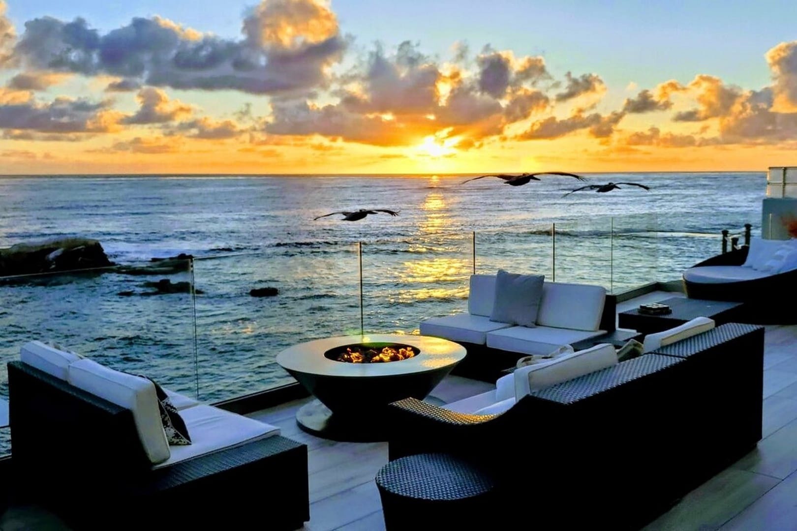 Sea Glass in La Jolla, San Diego, has an oceanfront terrace with cushioned seating around a round fire table. Birds fly above the rocky shoreline as the sunset casts golden light across the water.