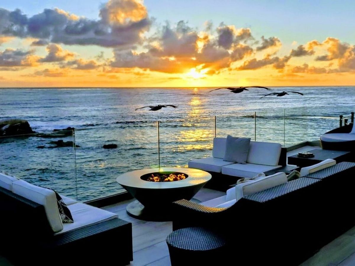 Sea Glass in La Jolla, San Diego, has an oceanfront terrace with cushioned seating around a round fire table. Birds fly above the rocky shoreline as the sunset casts golden light across the water.