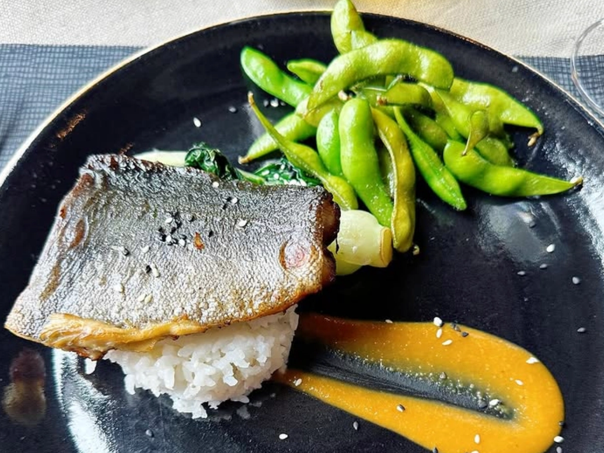 A plated dish featuring seared fish over white rice with sautéed greens, served alongside edamame and a streak of golden sauce.
