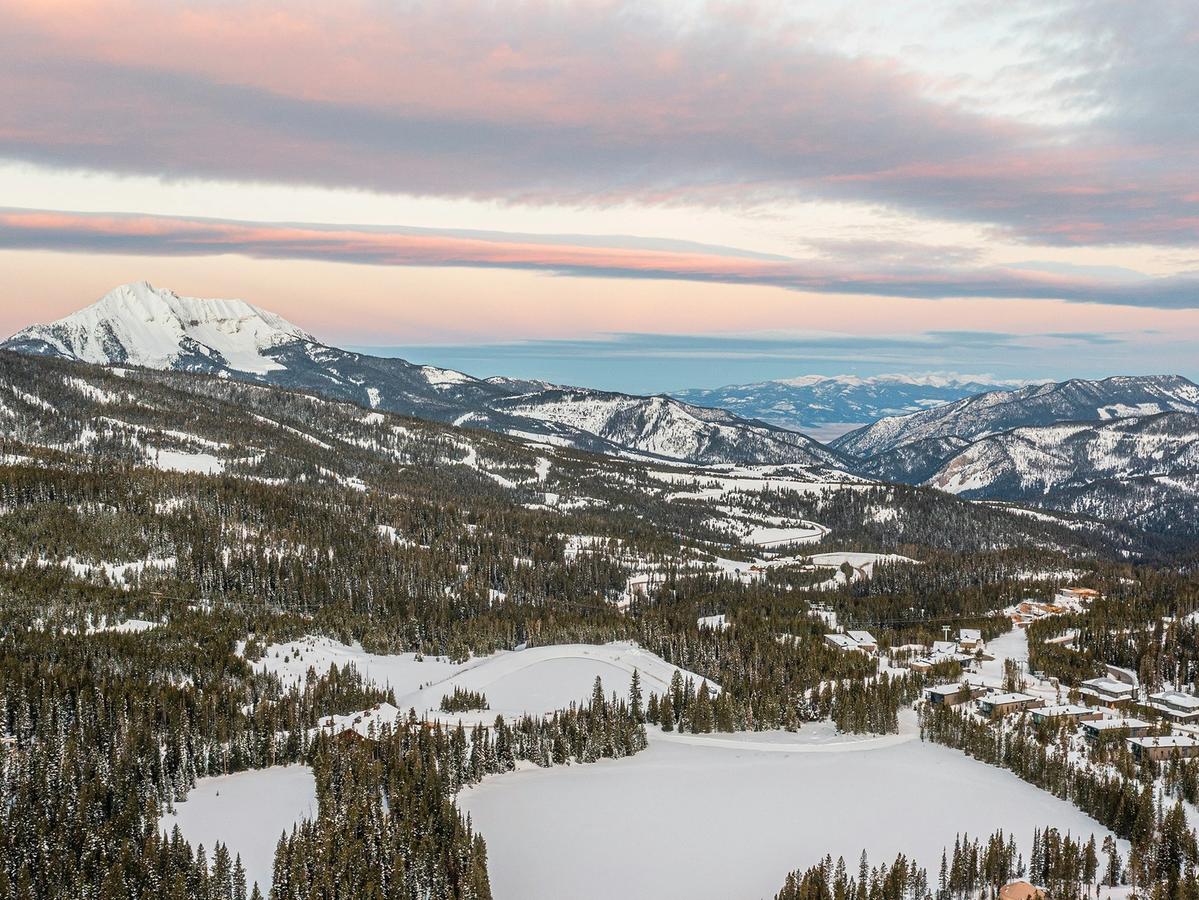 A majestic alpine landscape stretches beneath a pastel winter sky, with snow-draped forests and ski slopes leading toward a dramatic, isolated peak—likely Lone Mountain in Big Sky, Montana.