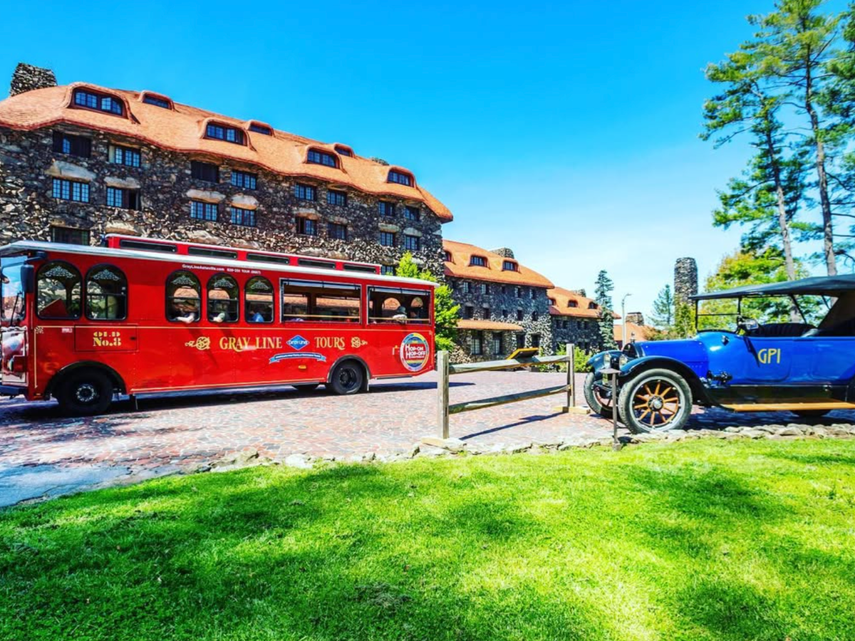 A bright red Gray Line Tours trolley and a vintage blue car sit in front of the historic stone Grove Park Inn under a clear blue sky.