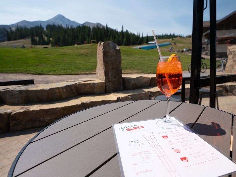 A bright orange drink sits on a table with a menu labeled “après ski” and a view of grassy slopes and mountains beyond. It’s the perfect spot to relax after a day of outdoor fun in Big Sky.