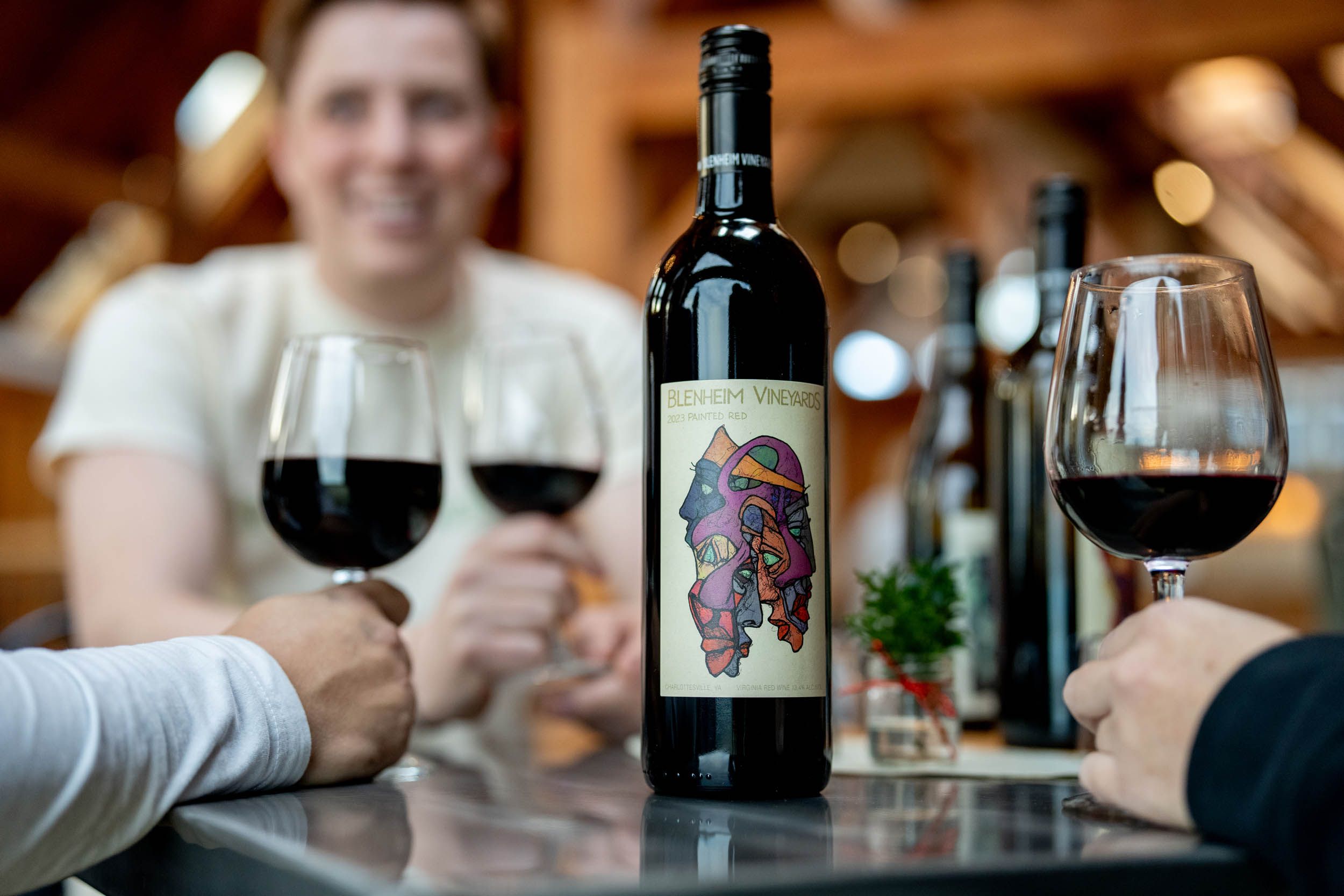 A bottle of Blenheim Vineyards red wine stands center stage as guests enjoy glasses of wine inside a rustic tasting room. The cozy indoor setting and cheerful atmosphere show the vineyard’s blend of art, community, and flavor.