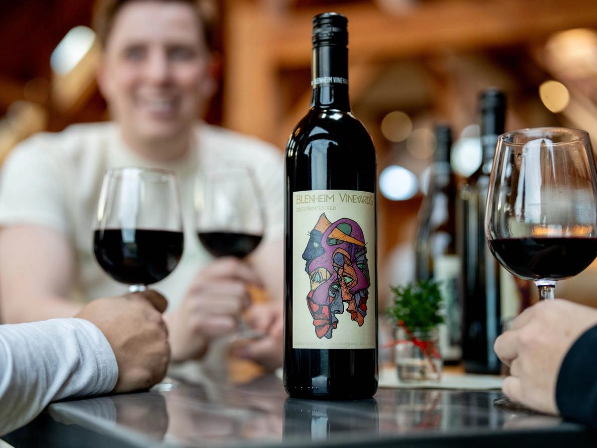 A bottle of Blenheim Vineyards red wine stands center stage as guests enjoy glasses of wine inside a rustic tasting room. The cozy indoor setting and cheerful atmosphere show the vineyard’s blend of art, community, and flavor.