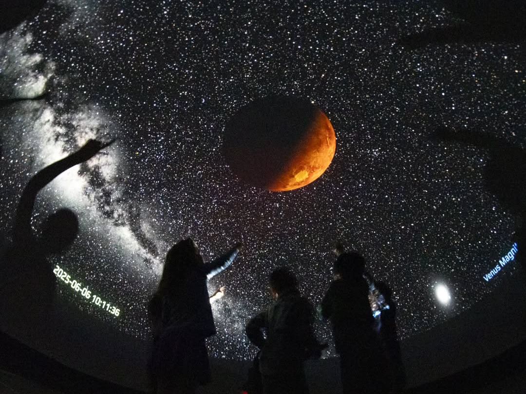 Visitors point toward a dazzling projection of the night sky inside a planetarium, with a glowing moon and Milky Way stretching across the dome. A captivating experience at the Oregon Observatory.