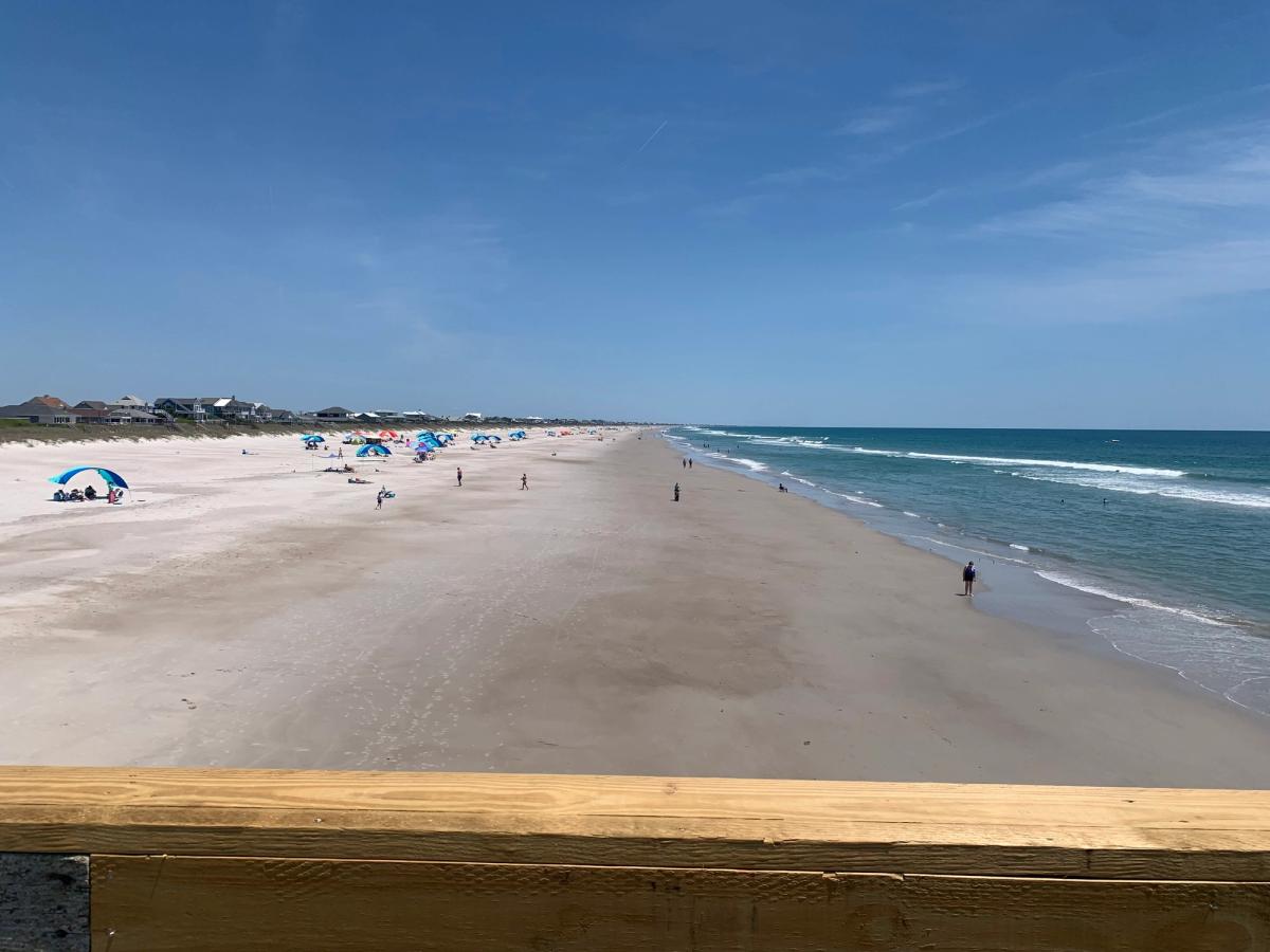 A sweeping view from the Jolly Roger Inn and Pier looks out over a wide stretch of white sand beach dotted with colorful umbrellas and beachgoers along the Carolina shoreline. Gentle turquoise waves roll in from the Atlantic under a clear blue sky, with beach cottages lining the dunes in the distance.