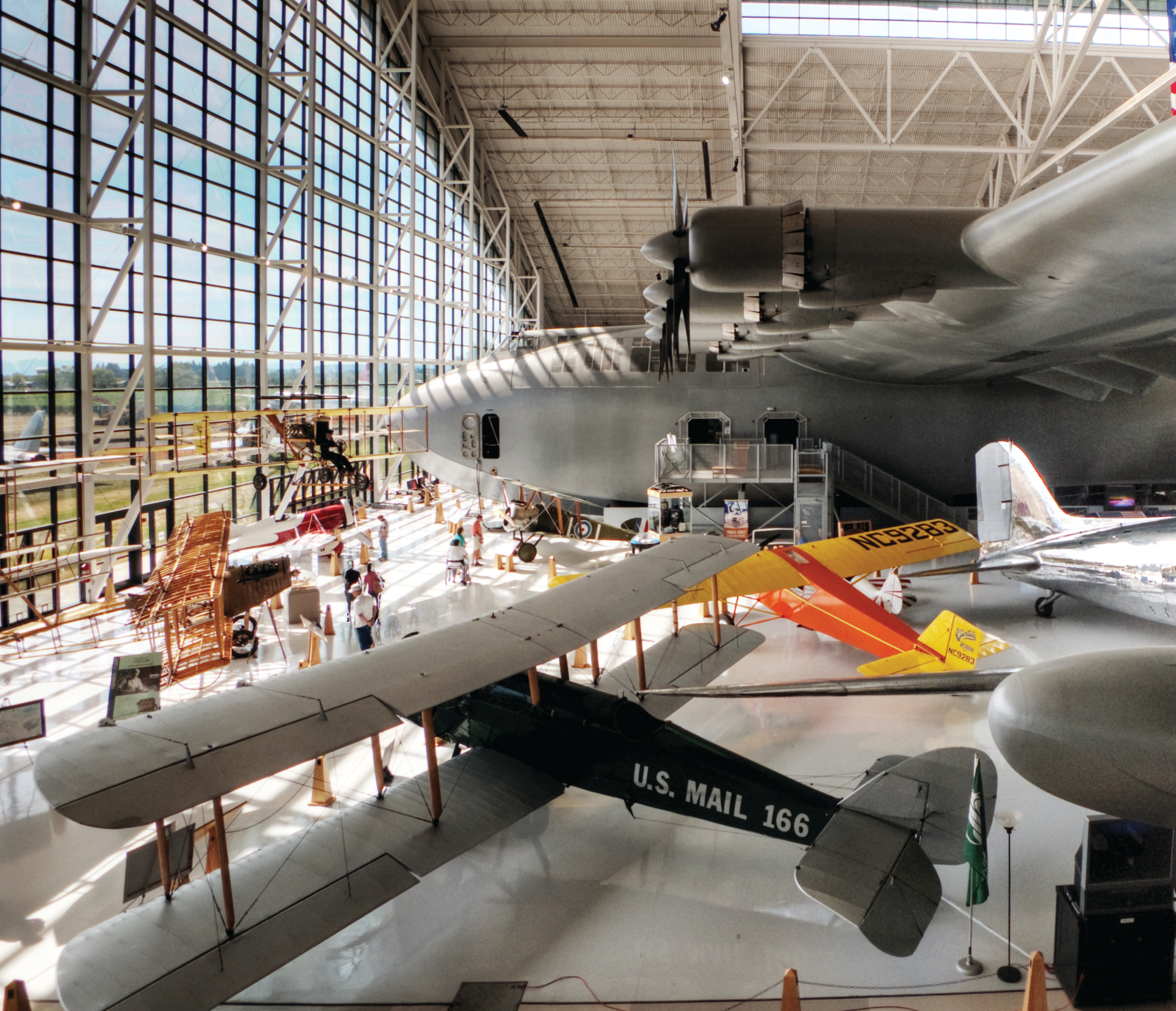 Historic airplanes fill a bright hangar at the Evergreen Aviation and Space Museum, including the famous Spruce Goose. Large glass windows shine natural light across the aircraft and visitors exploring the exhibits. The space feels open, modern, and full of aviation history.