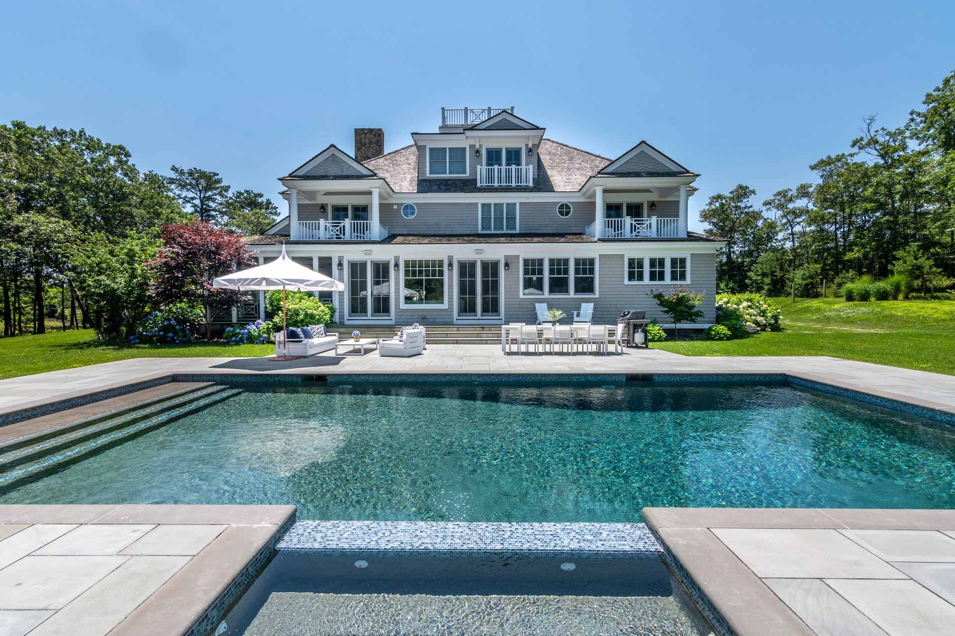 This bright backyard pool sits in front of a beautiful coastal-style home surrounded by green lawns and trees. The outdoor seating, umbrella, and dining area make it feel perfect for easy summer days spent outside. The whole scene gives a clean, calm, and welcoming vibe.