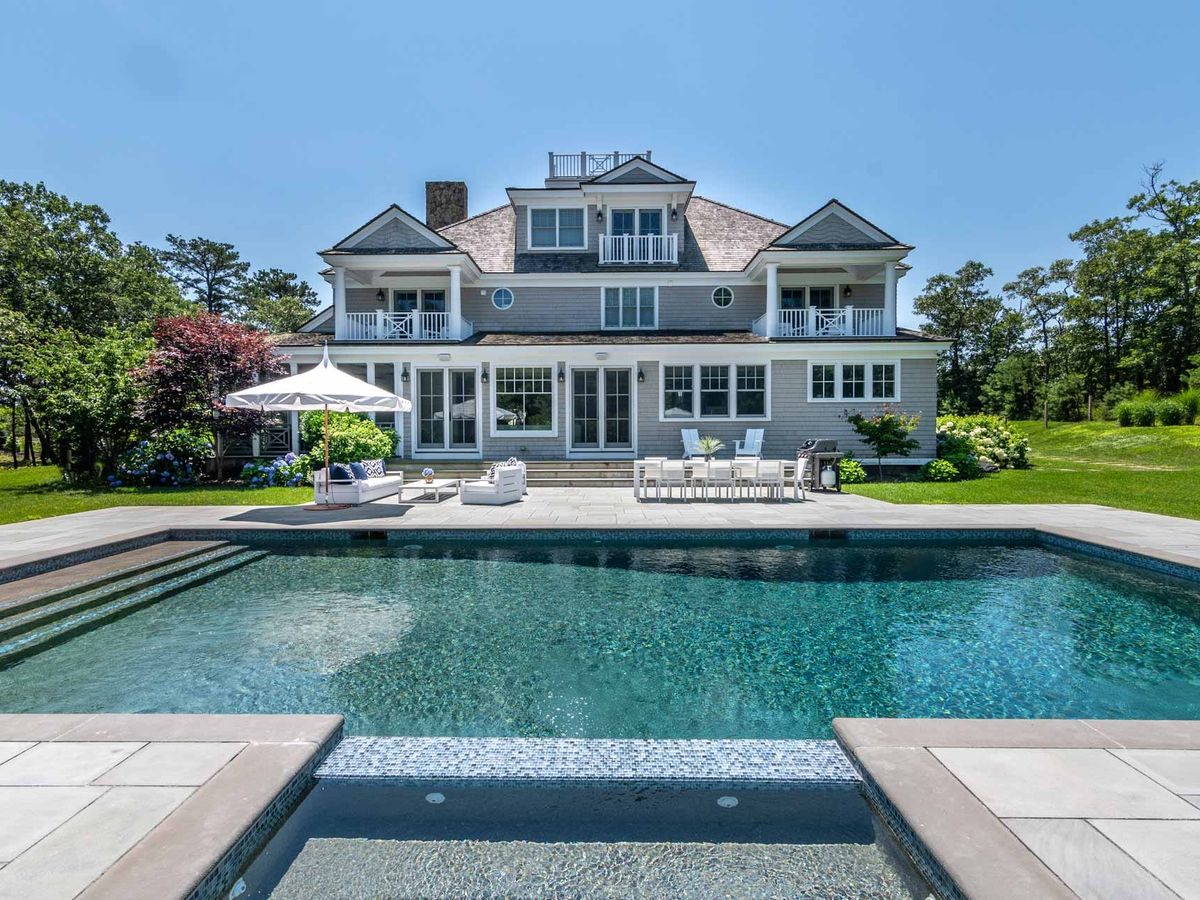 This bright backyard pool sits in front of a beautiful coastal-style home surrounded by green lawns and trees. The outdoor seating, umbrella, and dining area make it feel perfect for easy summer days spent outside. The whole scene gives a clean, calm, and welcoming vibe.