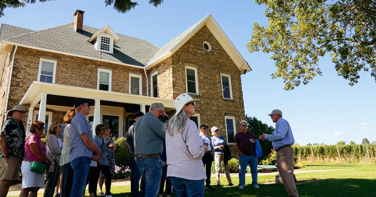A guided wine tour takes place on the lawn at Dr. Konstantin Frank Winery, with vineyard rows and a historic stone building behind. The outdoor setting adds to the classic Finger Lakes wine experience.