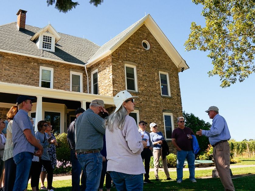 A guided wine tour takes place on the lawn at Dr. Konstantin Frank Winery, with vineyard rows and a historic stone building behind. The outdoor setting adds to the classic Finger Lakes wine experience.