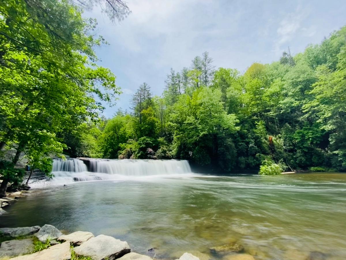 A wide, gentle waterfall flows into a calm, clear river surrounded by lush green trees and a rocky shoreline under a sunny sky.