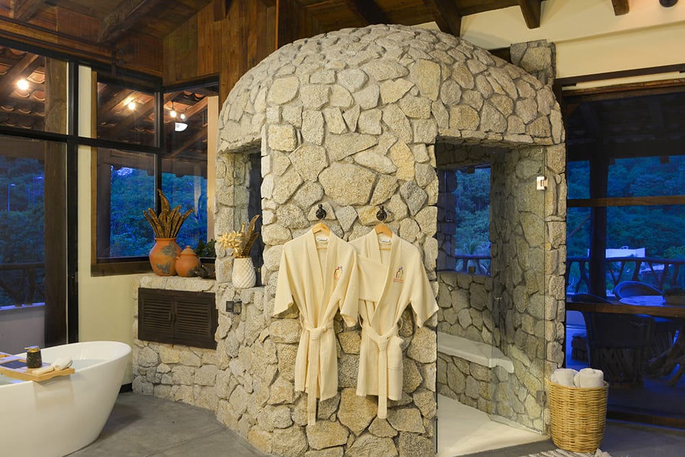 Luxury stone shower and soaking tub inside a spa-style bathroom in Huatulco, featuring rustic rock walls and soft warm lighting. This relaxing bathroom design gives you a peaceful retreat feel, perfect for a luxury vacation stay in Huatulco.