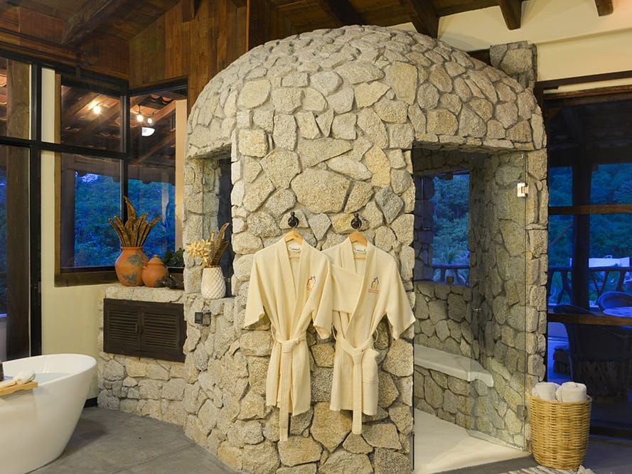Luxury stone shower and soaking tub inside a spa-style bathroom in Huatulco, featuring rustic rock walls and soft warm lighting. This relaxing bathroom design gives you a peaceful retreat feel, perfect for a luxury vacation stay in Huatulco.