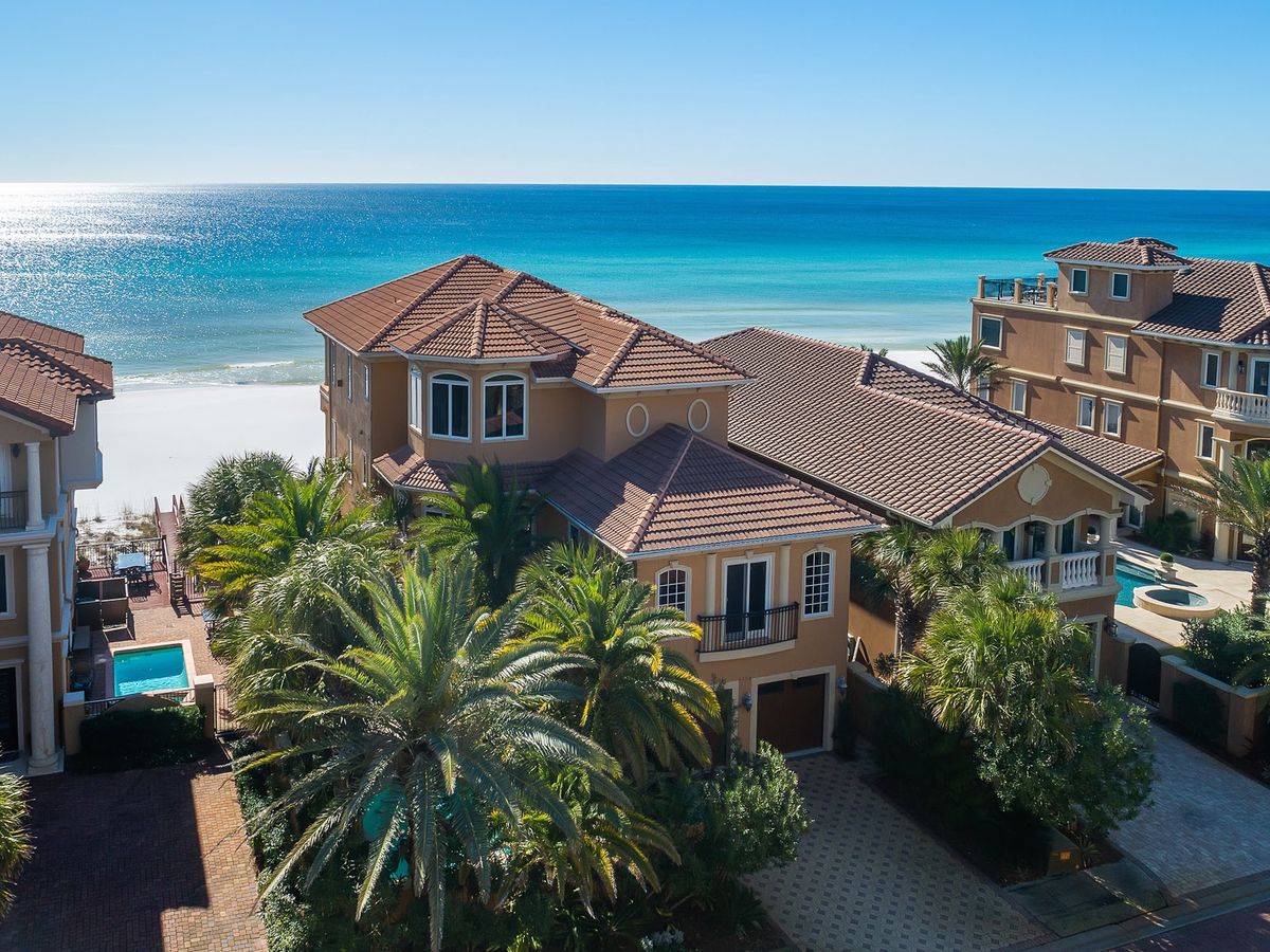 Mediterranean-style beachfront villas with terracotta roofs overlook a pristine stretch of white sand and turquoise ocean under a clear blue sky.