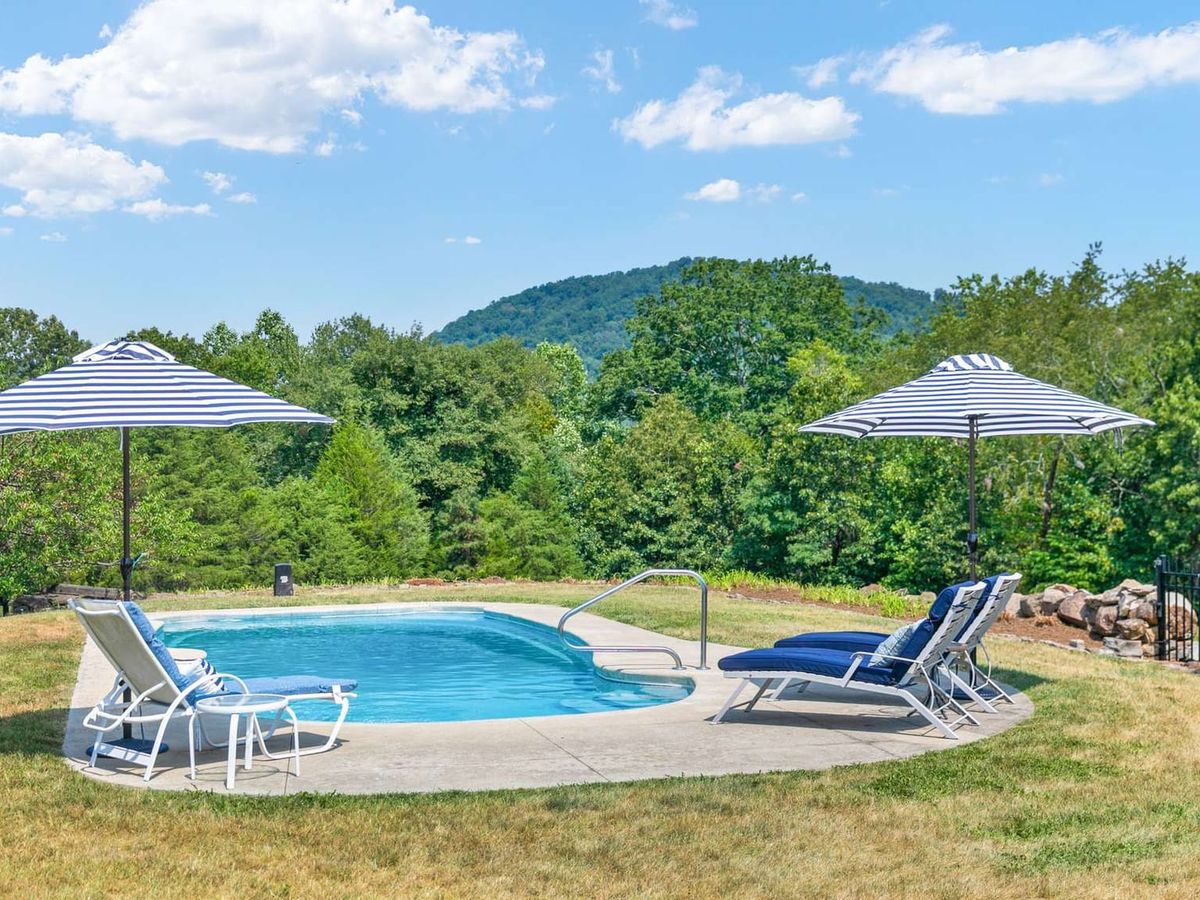 A kidney-shaped pool sits at the center of an open lawn at River Meadows Farm, framed by navy-striped umbrellas and lounge chairs with a forested hillside rising behind it. The mountain backdrop gives the setting a serene, secluded feel on a clear summer day.