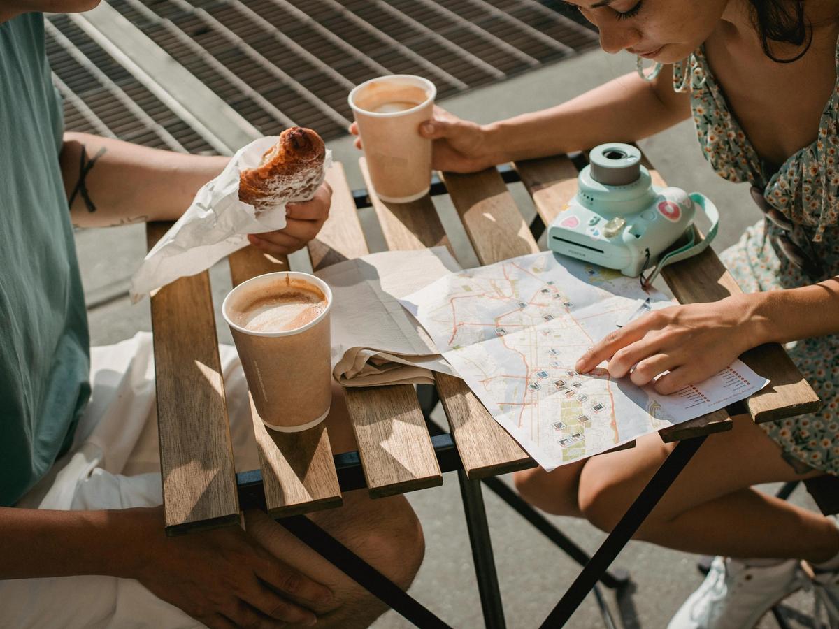 A couple sits at an outdoor café table with coffee, a pastry, and a map spread out as they plan their adventure. A camera rests nearby, capturing the essence of travel, exploration, and candid photography moments.