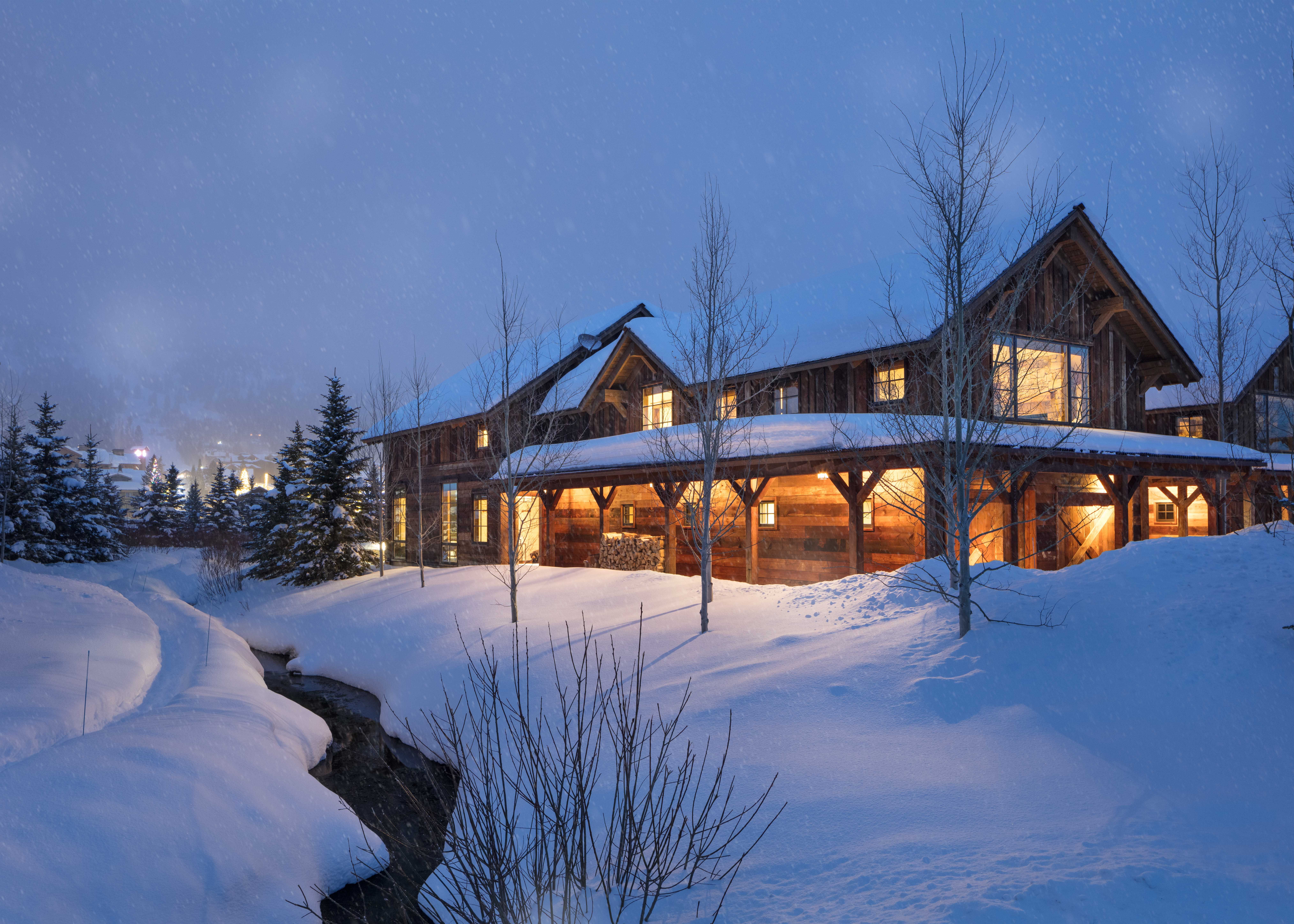Rustic mountain lodge warmly lit from within, surrounded by deep snowbanks and a winding creek during a gentle snowfall at twilight.