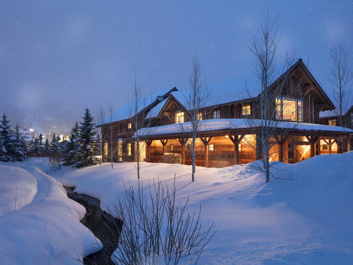 Rustic mountain lodge warmly lit from within, surrounded by deep snowbanks and a winding creek during a gentle snowfall at twilight.