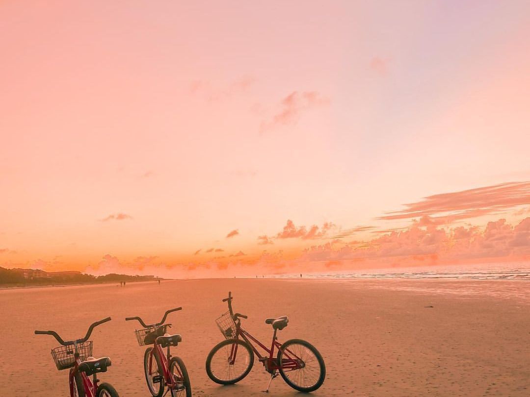 Three red bikes parked on a quiet beach at sunset, with soft pink and orange skies painting a dreamy scene—perfect for a weekend ride by the sea. A peaceful moment captured at golden hour.