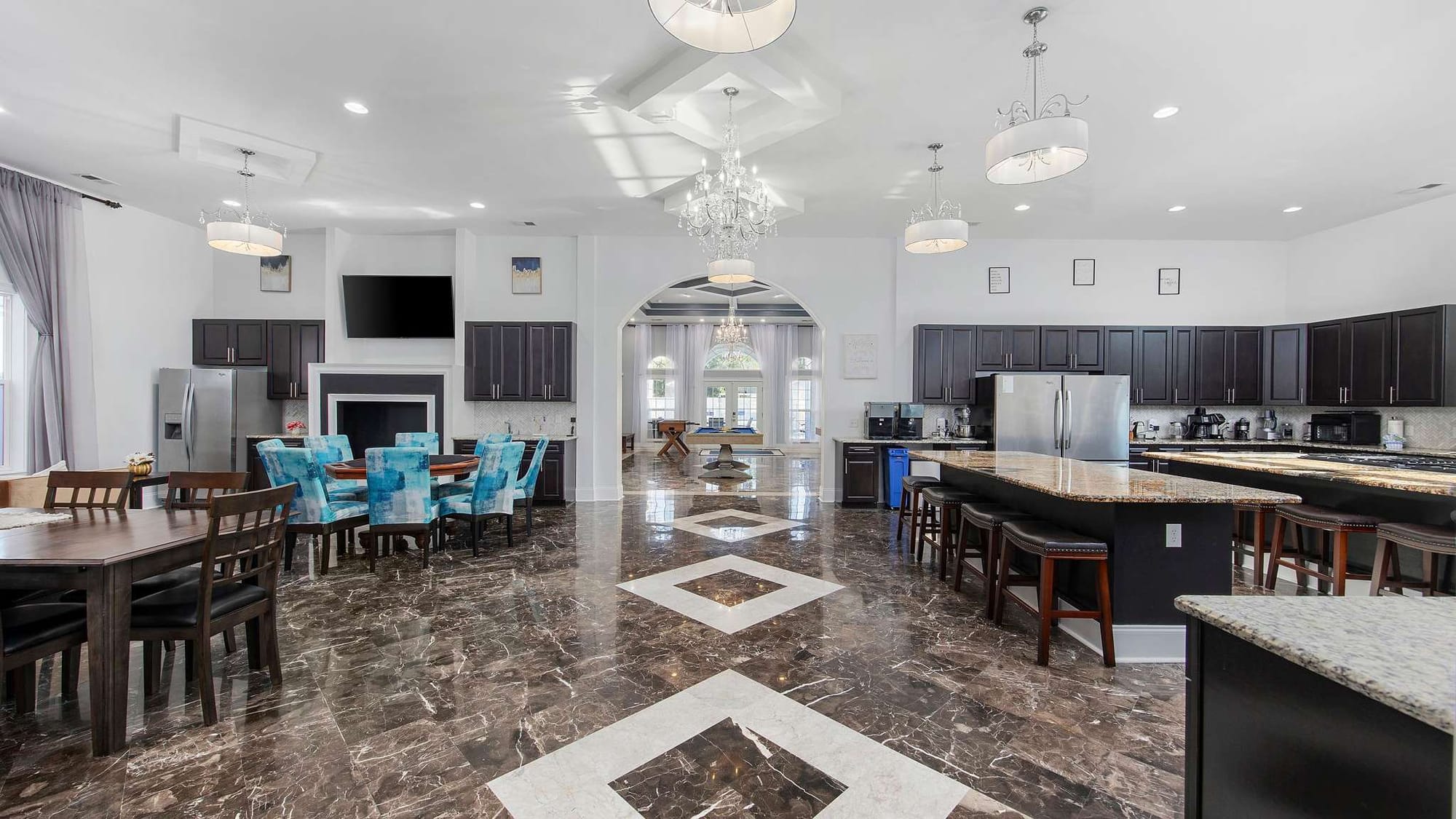 he grand open-plan living and kitchen area of Blue Paradise features polished marble floors with a geometric inlay pattern, a crystal chandelier, and multiple dining areas with teal upholstered chairs. A large arched doorway leads through to a game room and French doors beyond, while a fully equipped gourmet kitchen with granite countertops and dark cabinetry lines the right side of the space. 