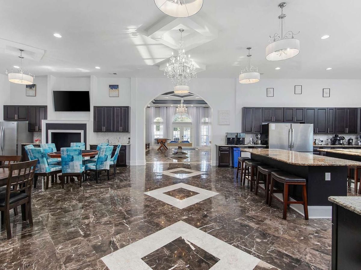 he grand open-plan living and kitchen area of Blue Paradise features polished marble floors with a geometric inlay pattern, a crystal chandelier, and multiple dining areas with teal upholstered chairs. A large arched doorway leads through to a game room and French doors beyond, while a fully equipped gourmet kitchen with granite countertops and dark cabinetry lines the right side of the space.
